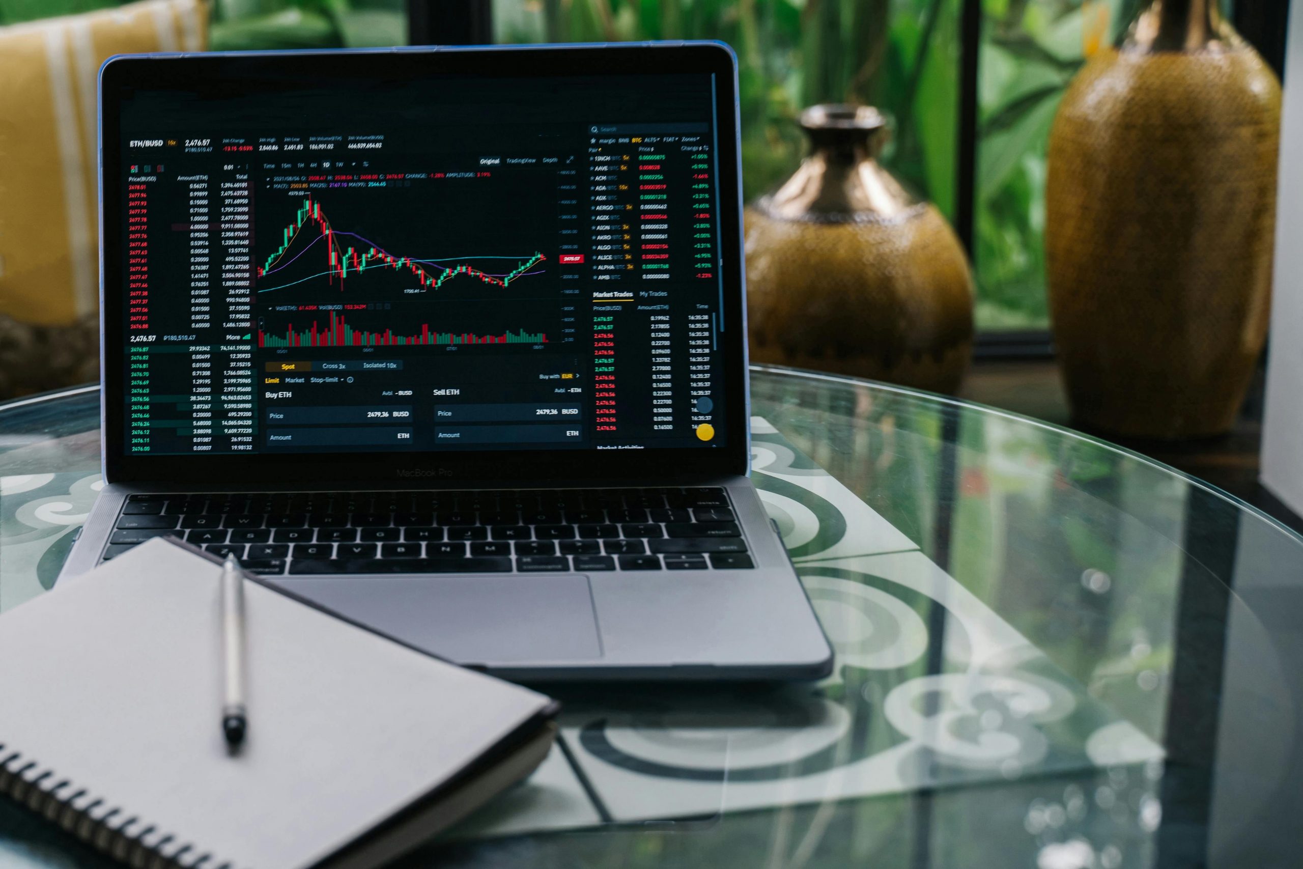 A laptop sitting on a table. The screen displays stocks in graph form. A blank journal sits on the laptop.