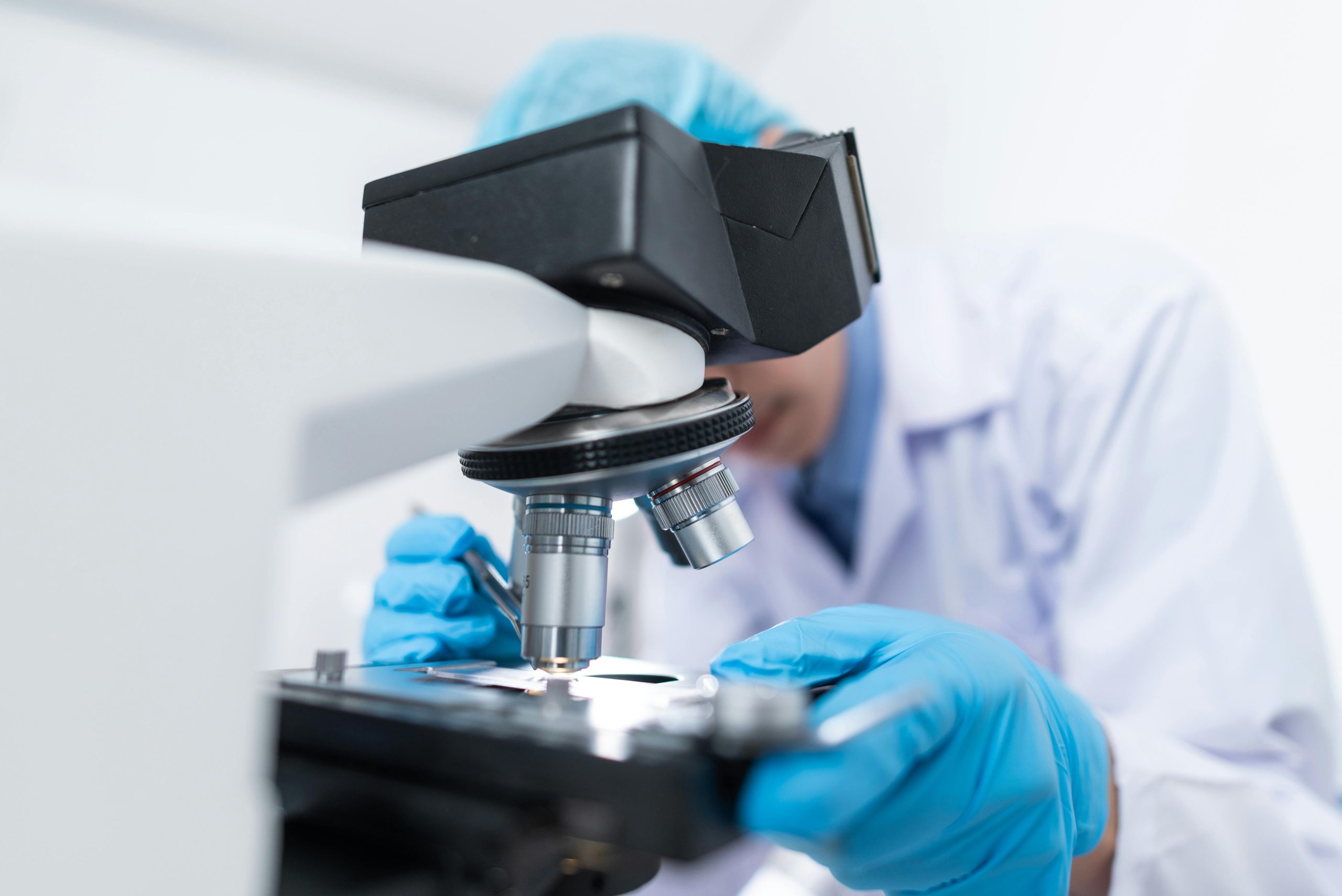 A person in a labcoat and gloves looks into a microscope.
