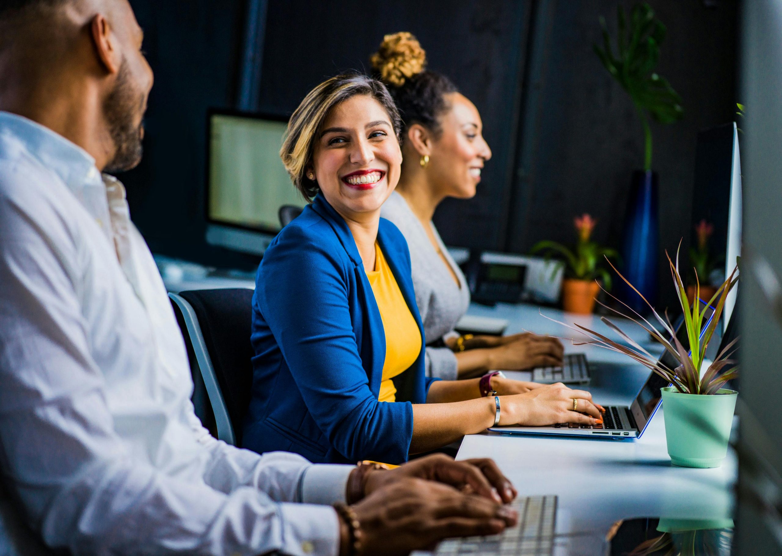 A woman smiles at the camera. On her left is a man, and her right is a woman. They are all typing at computers.