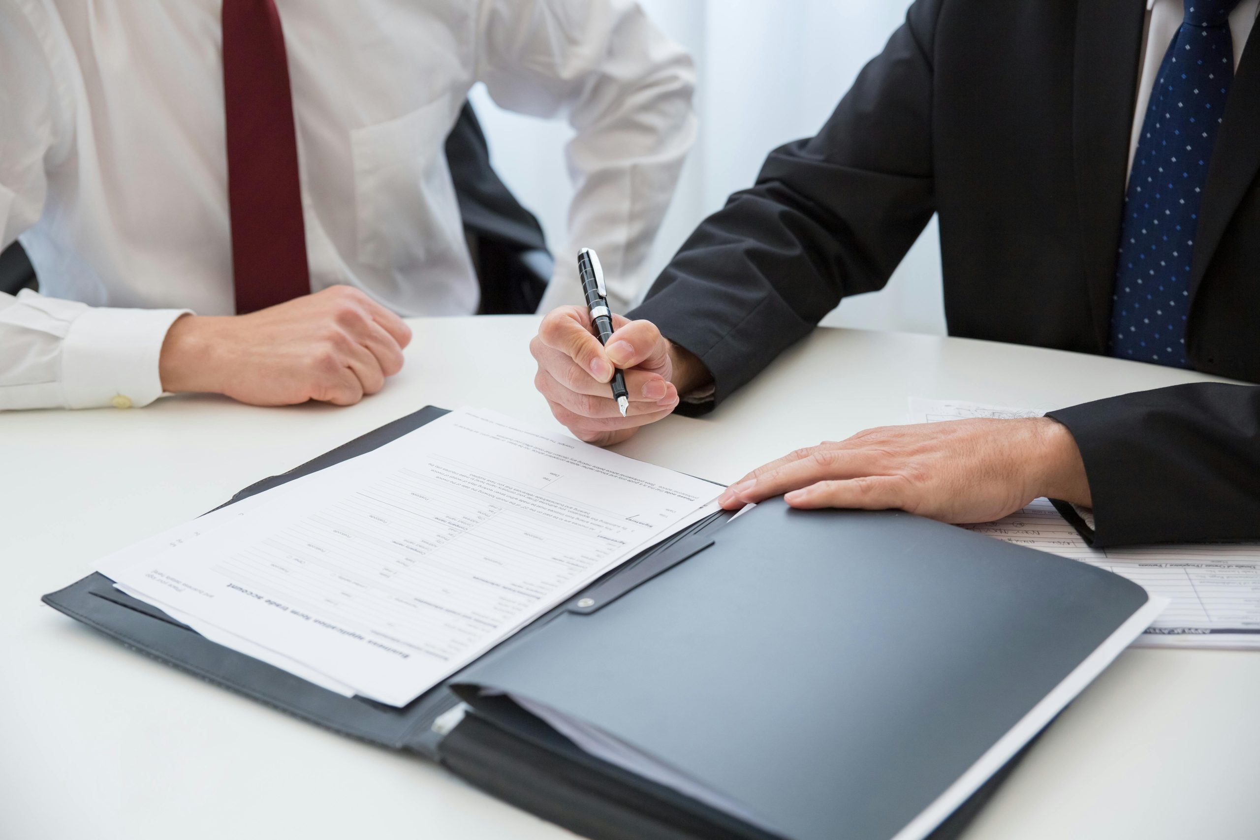 Two people sit in front of a folio of papers. The person on the right is signing a document.