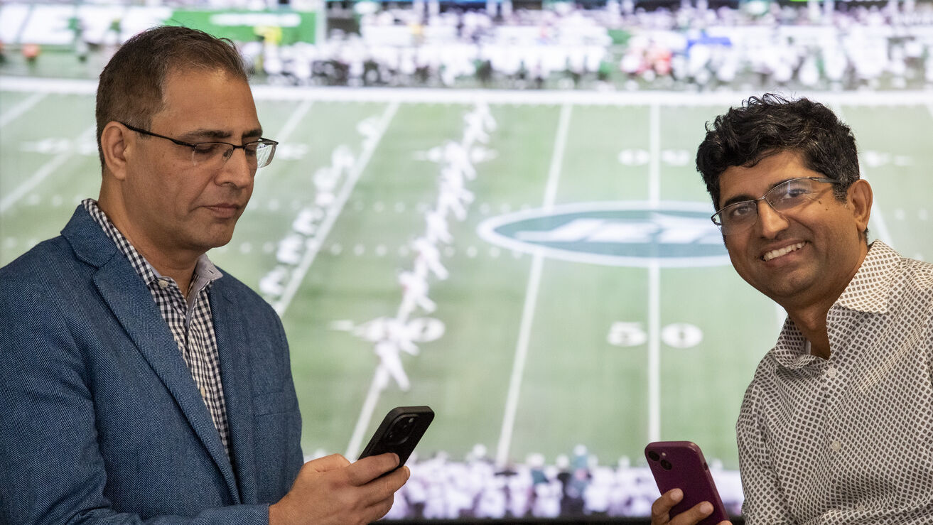 Two men stand in front of a football game. One is looking at his phone, while the other is looking towards the viewer.