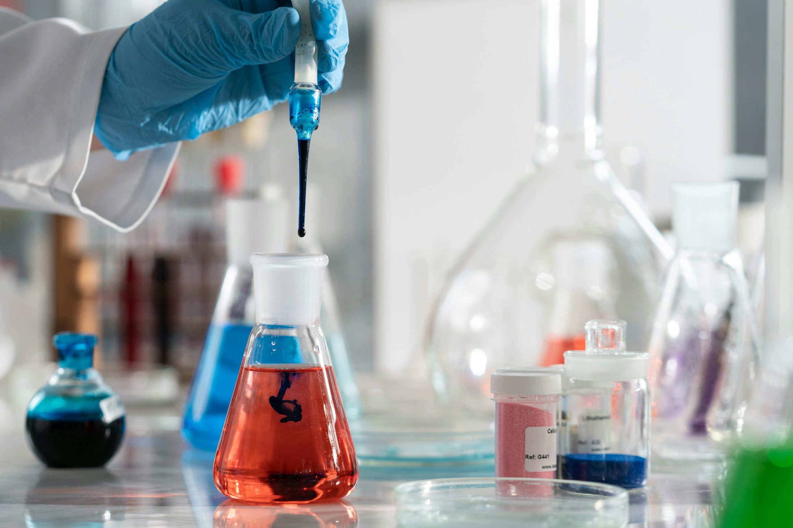 A gloved hand puts a liquid into a beaker. Multiple beakers are in the background.