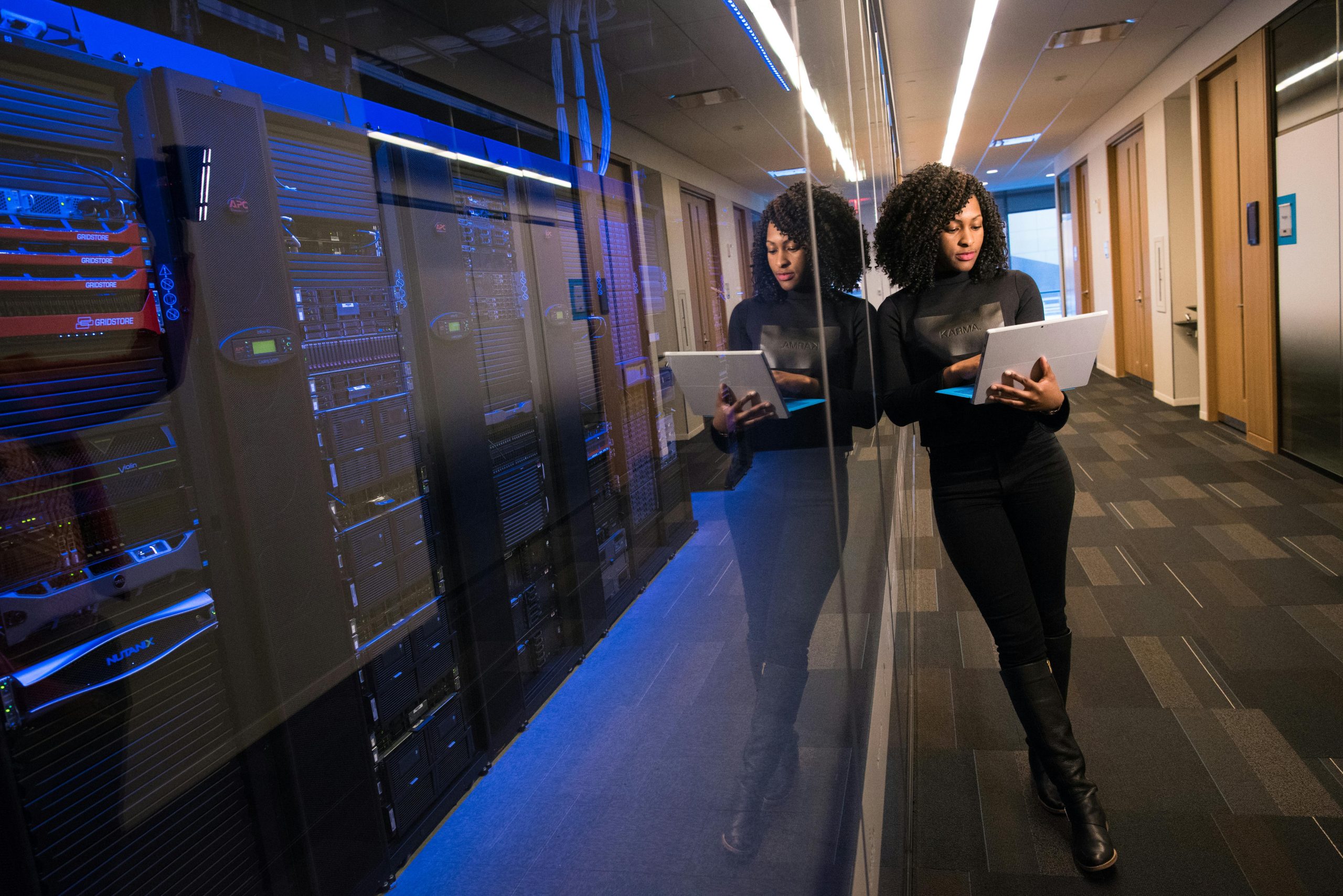 A woman leans against a glass wall, behind which are rows of servers.