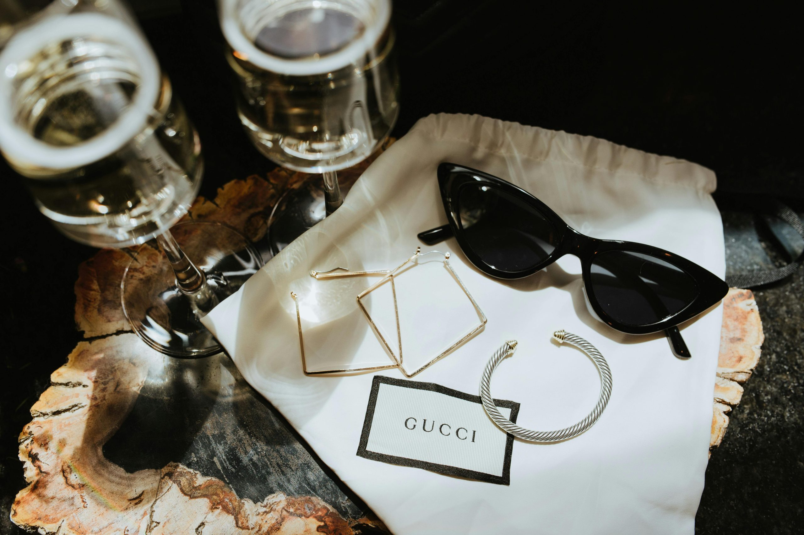 Earrings, a bracelet and sunglasses sit on a bag with a Gucci label. Two champagne glasses sit on the same table.