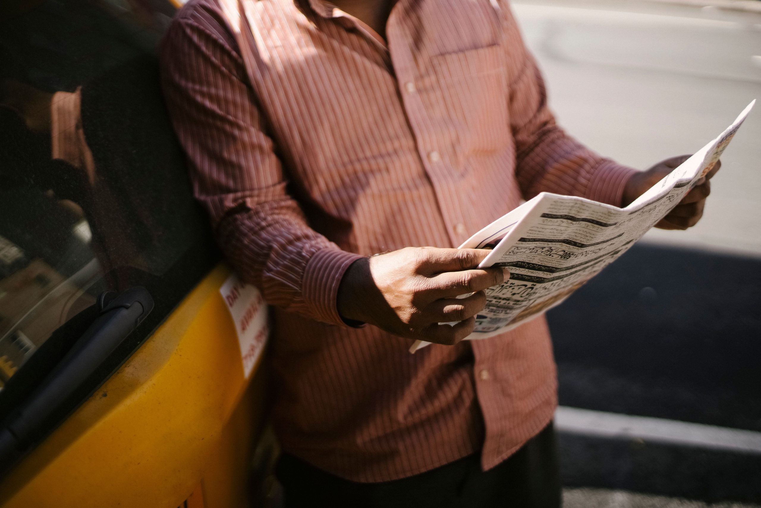A person holds a newspaper and leans against a car.